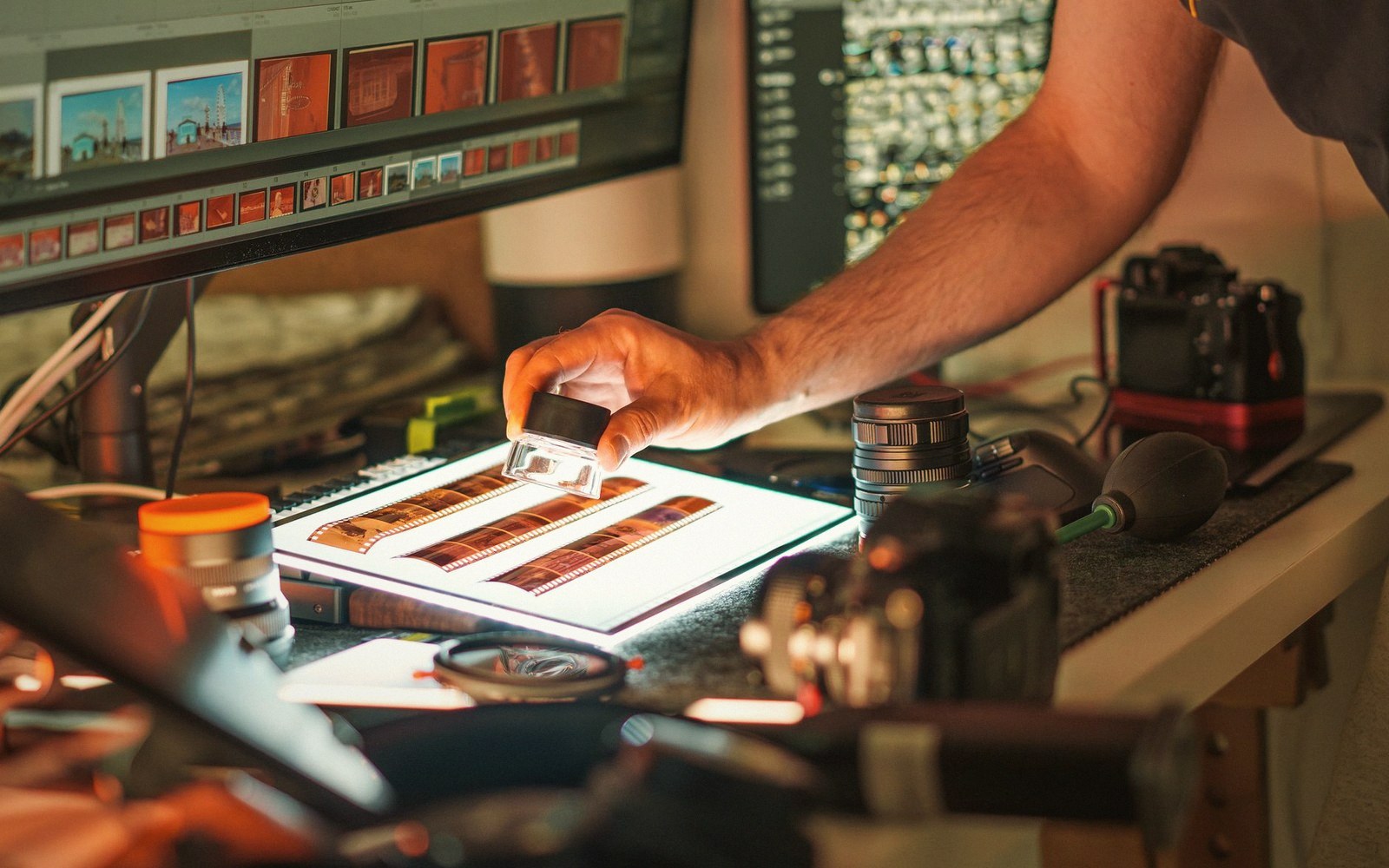 A person examines film negatives with a loupe on a lightbox beside camera equipment and computer screens