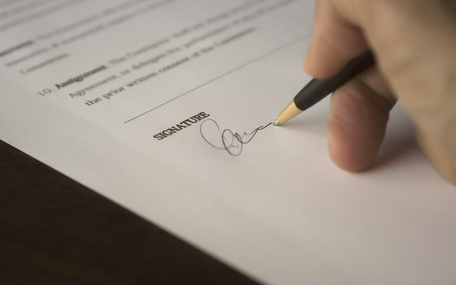 A hand signs a document on the line marked "signature" with a pen